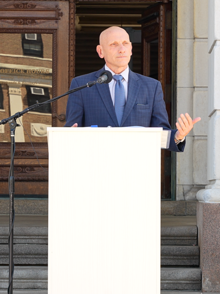 A man in a navy plaid suit and blue tie speaks at an outdoor podium on the steps of the Rhode Island State House, gesturing with one hand. A microphone on a stand is positioned in front of him.