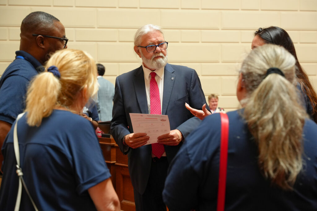 A legislator in a navy suit and red tie listens attentively while a group of advocates in blue CPNRI shirts speaks with him inside the State House. He holds an advocacy one-pager.