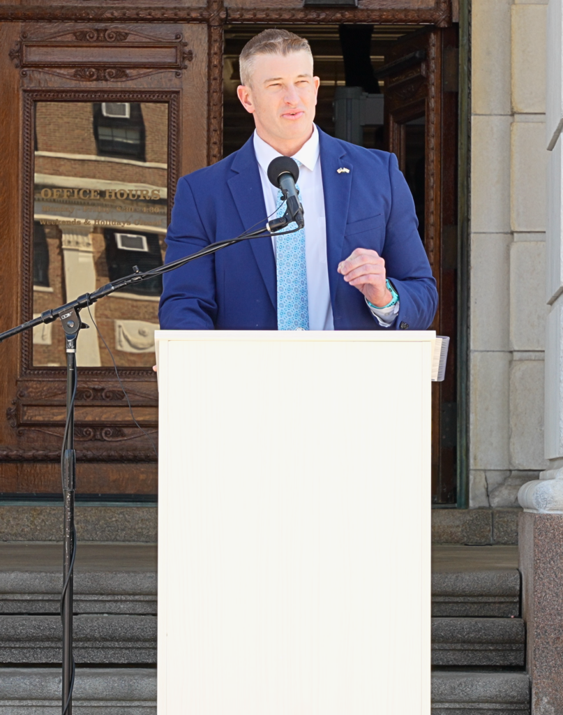 A man in a blue suit and light blue patterned tie speaks at an outdoor podium on the steps of the Rhode Island State House. A small American flag pin is visible on his lapel.