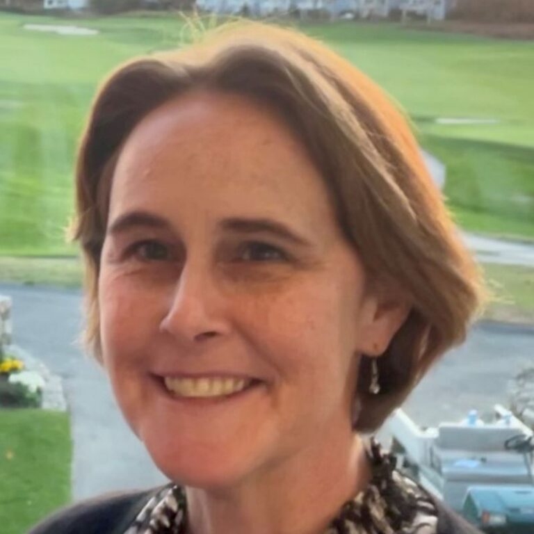 A woman with shoulder-length reddish-brown hair smiles warmly at the camera, showing her teeth, with dangling earrings visible. She wears a black and white patterned top. The background shows a sunlit golf course with green fairways, a paved driveway, and a parked RV or camper visible to the right.