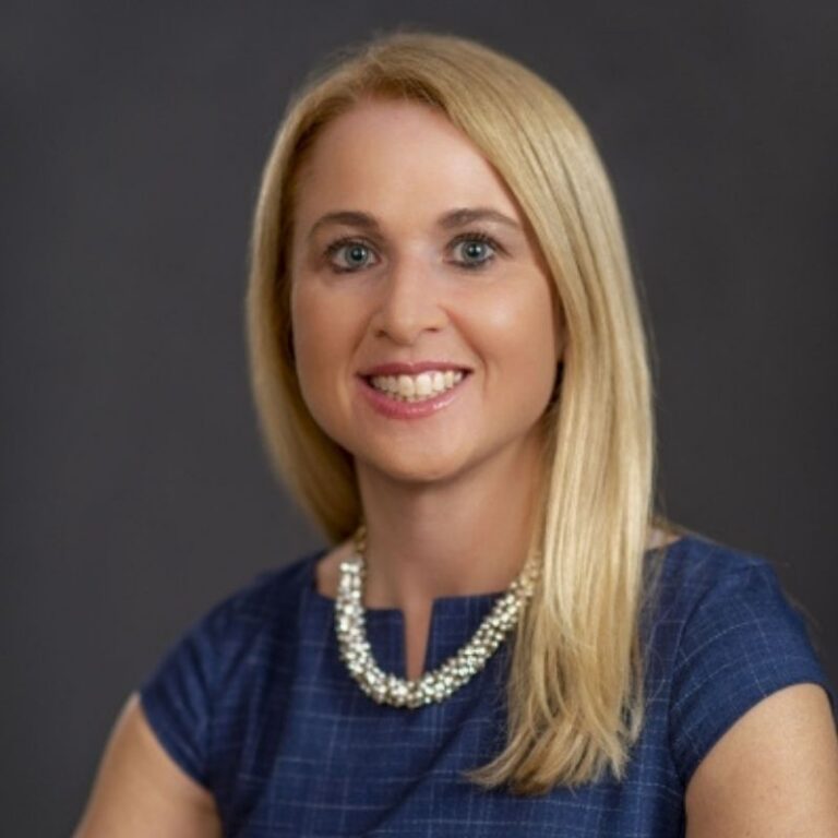 Beth Bixby smiles warmly at the camera in a professional headshot, wearing a navy checked top and a chunky pearl statement necklace. She has long, straight blonde hair and is photographed against a solid dark gray background.