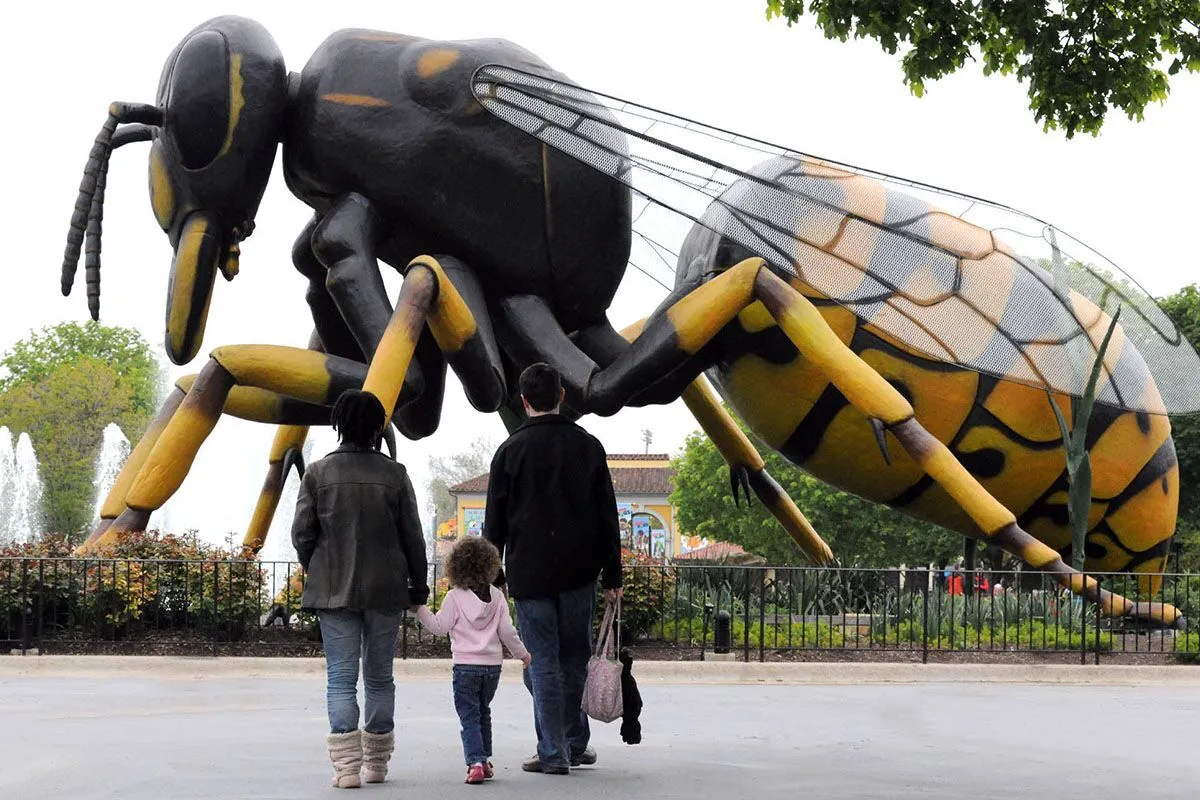 A family with a young child walks toward a massive, realistic sculpture of a wasp in an outdoor park setting. The oversized insect towers above them, with detailed wings and yellow-and-black coloring, creating a dramatic sense of scale. Trees, a fence, and landscaped greenery surround the scene.