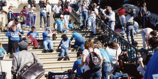 Disability rights activists crawl up the steps of the U.S. Capitol during the 1990 Capitol Crawl protest, while photographers and reporters document the moment. Many participants have left their wheelchairs and mobility devices behind at the bottom of the stairs, pulling themselves upward with their hands as part of a demonstration calling for passage of the Americans with Disabilities Act.