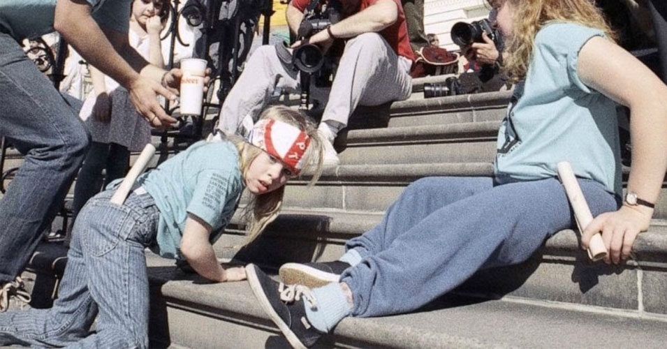 A young disability rights activist crawls up the marble steps of the U.S. Capitol during the 1990 Capitol Crawl protest, while photographers and supporters watch nearby. The child wears a red headband and blue shirt, pulling herself upward with her hands as part of the demonstration calling for passage of the Americans with Disabilities Act.