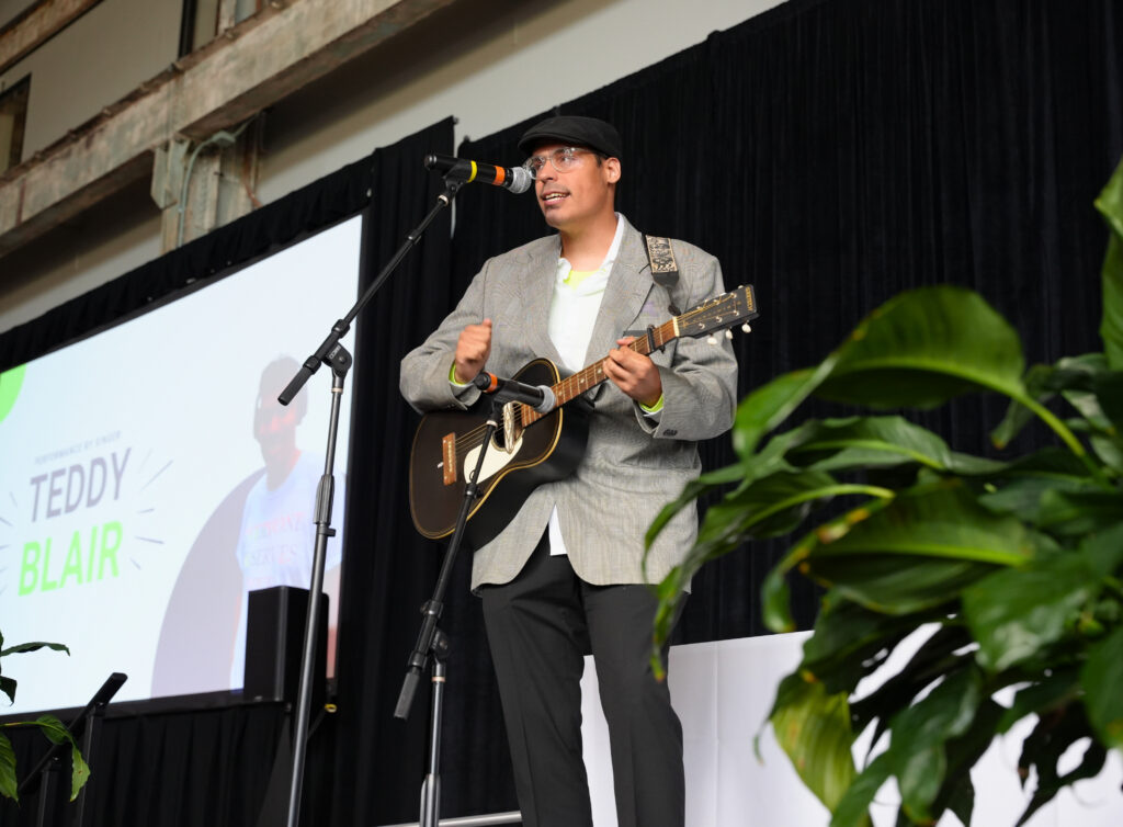 A singer in a gray plaid blazer, flat cap, and clear-framed glasses performs on stage, singing into a microphone while playing a black acoustic guitar. Behind him, a large screen displays his photo with the text Performance by Singer Teddy Blair, and leafy green plants frame the foreground.