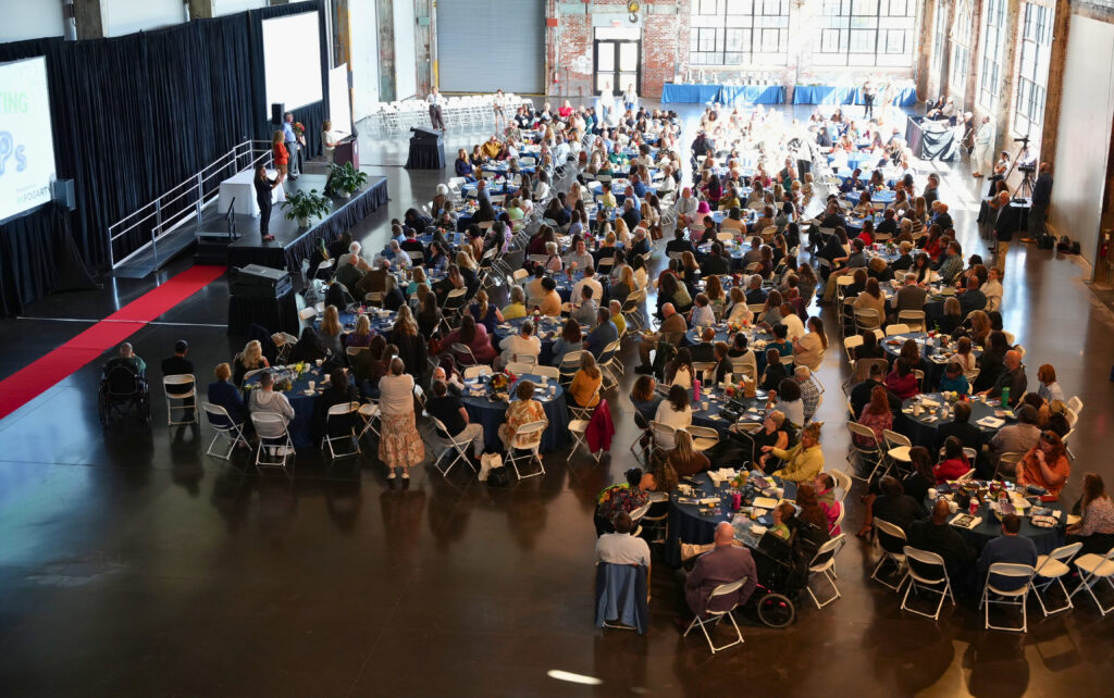 From a high vantage point, a large crowd seated at round navy-draped tables faces a black stage on the left, where a speaker addresses the room from a podium and a partial screen shows the Fogarty logo. Sunlight pours through tall industrial windows at the back of the hall, a red carpet runs beside the stage, and a camera operator films from the right side.