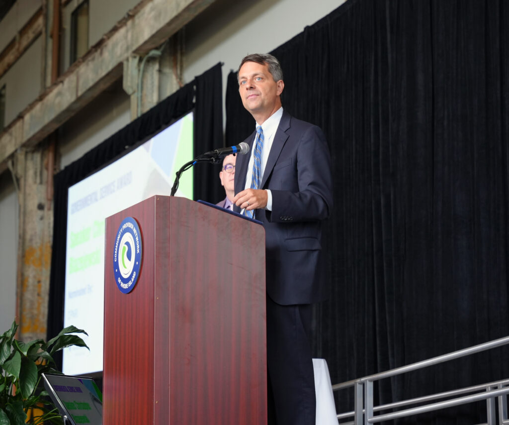 A man in a navy suit and blue striped tie stands at a wooden podium bearing the Community Provider Network of Rhode Island seal, looking out toward the audience. Behind him, a screen displays Governmental Service Award, nominated by CPNRI, and another man in glasses stands in the background.