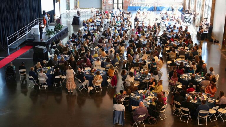 A high vantage point looks across a large converted industrial hall with brick walls and tall windows, where hundreds of attendees sit at round tables draped in navy blue cloths, each with small flower centerpieces, cups, and papers. On a raised black stage at the upper left, a speaker addresses the room from a podium while several people stand nearby, and a red carpet runs along the floor beside the stage. Two people using wheelchairs are seated among the tables in the foreground, and a videographer with a camera on a tripod records from the right side of the room as sunlight streams in through the windows.