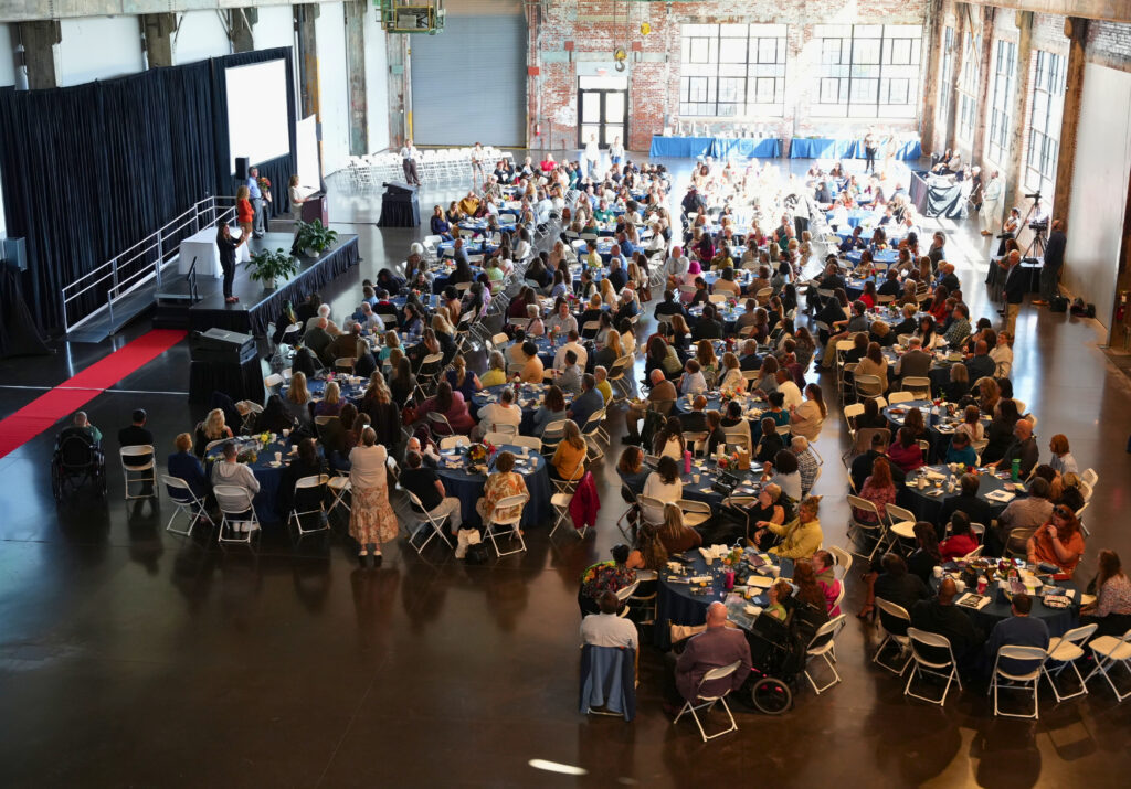 A high, wide view looks across a sunlit converted industrial hall with brick walls and tall factory windows, where hundreds of guests sit at round tables covered in navy cloths with flower centerpieces. On a black stage at the upper left, a speaker stands at a podium with several people nearby, a red carpet runs along the floor beside the stage, and a videographer films from a tripod on the right. Two guests using wheelchairs are seated among the tables in the foreground.