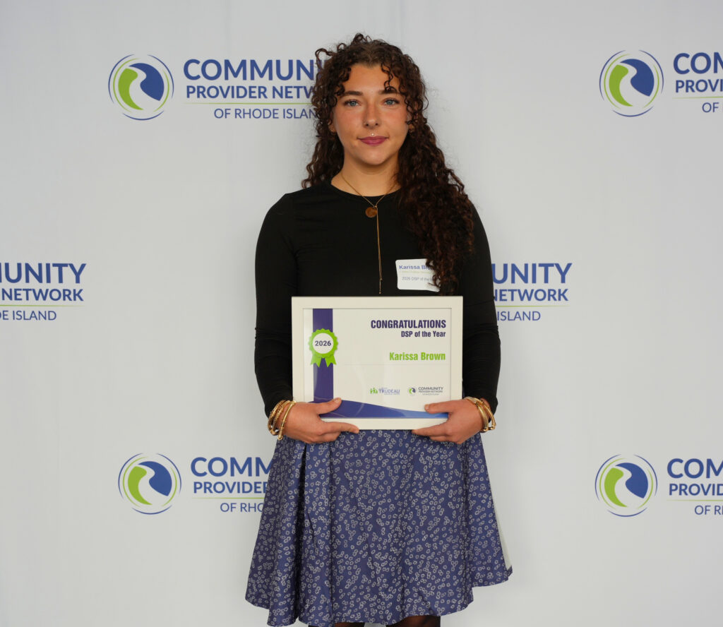 Karissa Brown, a young woman with long dark curly hair, stands in front of a Community Provider Network of Rhode Island step-and-repeat backdrop, holding a framed certificate that reads Congratulations, DSP of the Year, 2026, Karissa Brown. She wears a black long-sleeve top, a navy floral skirt, gold bangles, and a name tag, and gives a calm, slight smile.