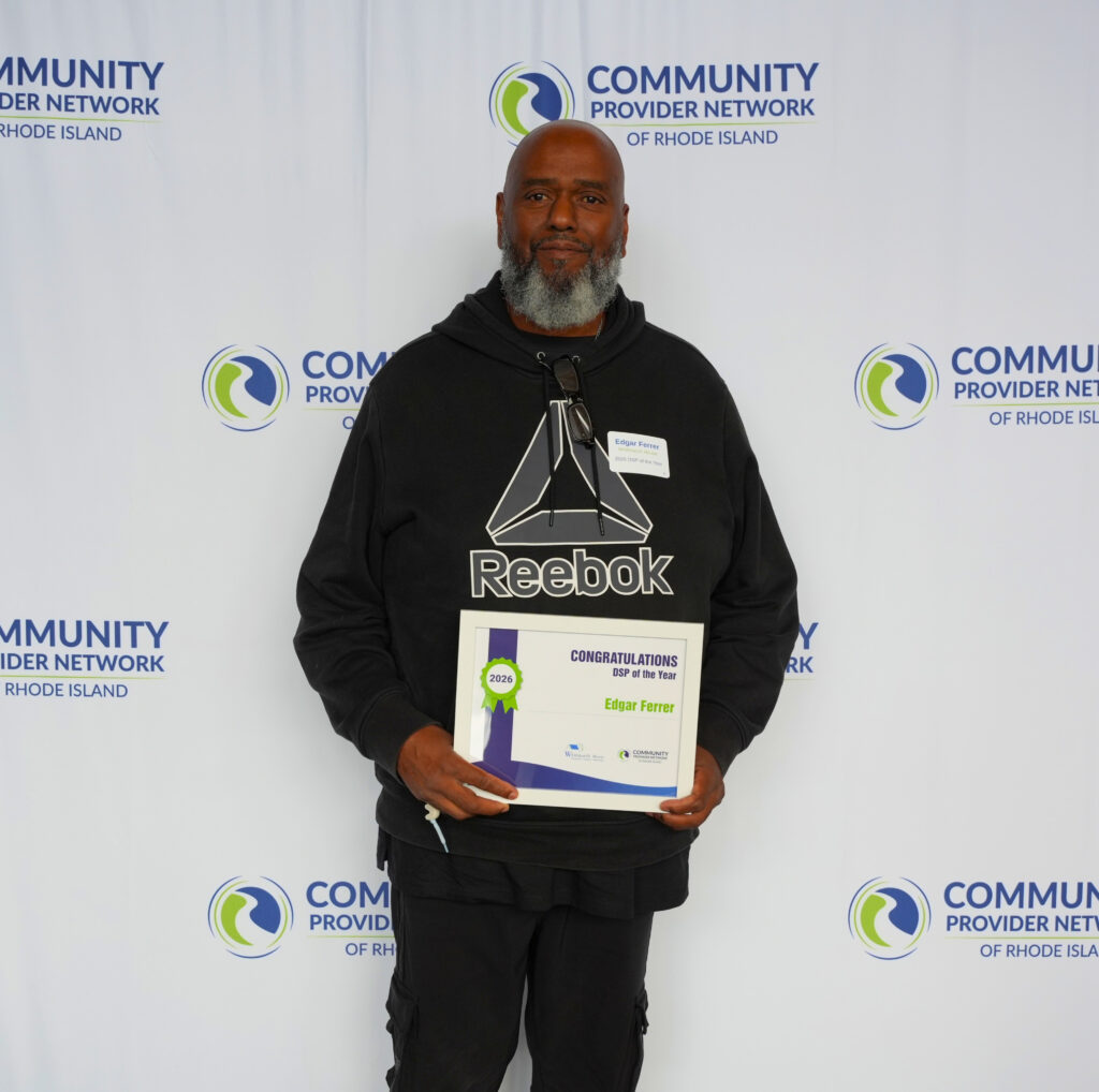 Edgar Ferrer, a man with a full gray beard, stands in front of a Community Provider Network of Rhode Island backdrop holding a framed certificate that reads Congratulations, DSP of the Year, 2026, Edgar Ferrer. He wears a black hoodie and pants, with glasses hooked on his collar and a name tag identifying him as a 2026 DSP of the Year from Whitmarsh House.