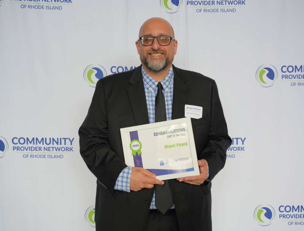 Shawn Picard, a smiling man with glasses and a salt-and-pepper beard, poses in front of a Community Provider Network of Rhode Island backdrop, pointing proudly at his framed certificate that reads Congratulations, DSP of the Year, 2026, Shawn Picard. He wears a black suit, blue plaid shirt, and black tie, with a name tag listing Justice Resource Institute.