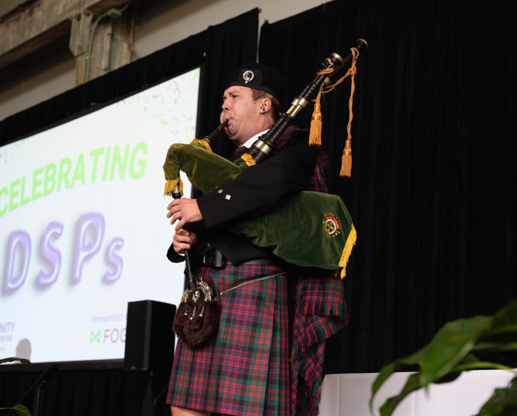 A bagpiper in full Highland dress plays on stage, wearing a red and green tartan kilt, a black jacket, a black cap with a silver badge, and a fur sporran, with a green velvet bag and gold tassels on his pipes. Behind him, a large screen reads Celebrating DSPs, with Presented by Fogarty partly visible at the bottom.