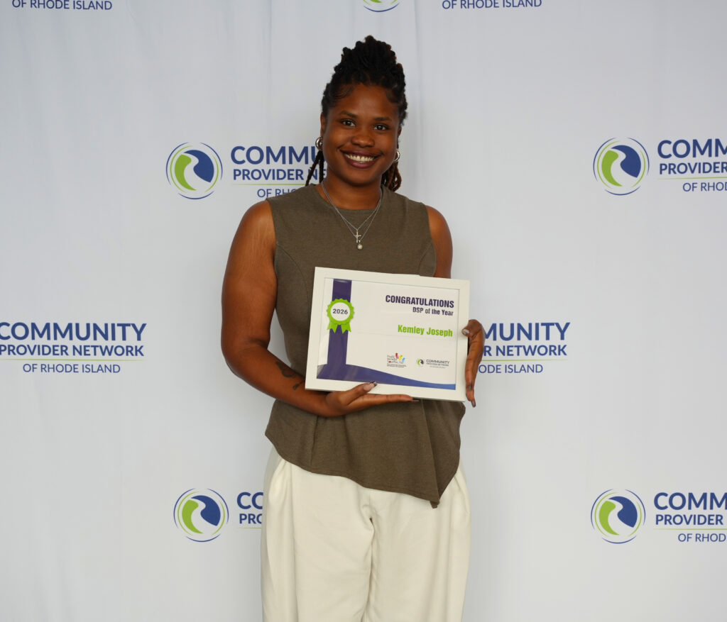Kemley Joseph, a smiling woman with her locs pulled up, stands in front of a Community Provider Network of Rhode Island backdrop, presenting her framed certificate that reads Congratulations, DSP of the Year, 2026, Kemley Joseph. She wears a sleeveless olive top, cream wide-leg pants, hoop earrings, and a delicate cross necklace.