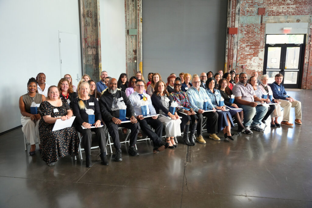 About 40 honorees sit and stand in several rows for a group photo inside a brick-walled industrial hall, in front of a large gray roll-up door. Most smile at the camera while holding certificate folders and small navy gift bags with white tissue paper in their laps.