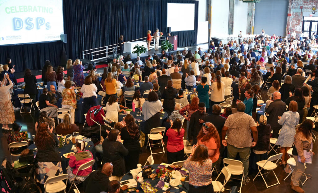 A packed room of guests rises to their feet in a standing ovation, clapping and cheering toward the stage, where several people stand near the podium. A large screen at the upper left reads Celebrating DSPs alongside the Community Provider Network of Rhode Island and Fogarty logos, and some guests hold up phones to capture the moment.