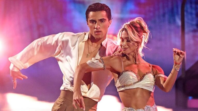Two ballroom dancers perform a dramatic pose on a stage washed in pink, purple, and blue light. On the left, a dark-haired man in an open, flowing white satin shirt, gold necklaces, and tan trousers extends one arm wide with an intense expression. In front of him, a woman with blonde hair in a loose updo smiles with her eyes closed, one arm curved upward and the other bent near her waist. She wears a white satin crop top and skirt trimmed with rhinestones, plus several sparkling bracelets.