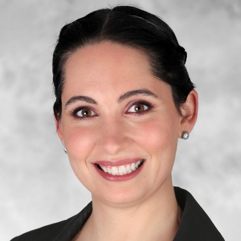 A woman with dark hair pulled back into a low updo smiles broadly against a light gray textured background. She wears a black jacket with sparkling stud earrings and defined makeup.