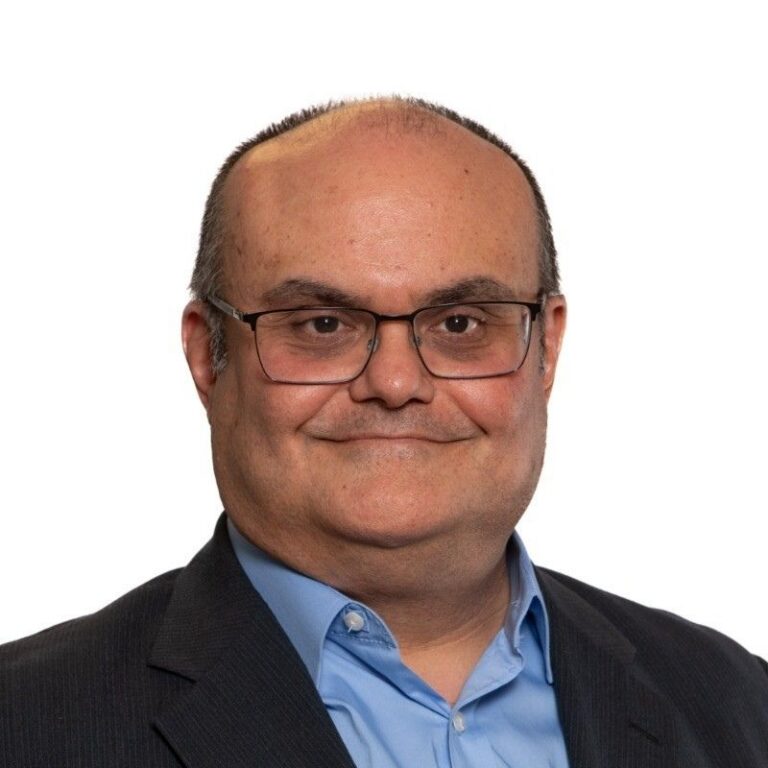 A man with short dark hair receding at the temples and rectangular wire-frame glasses smiles gently against a plain white background. He wears a dark suit jacket over a light blue button-down shirt.