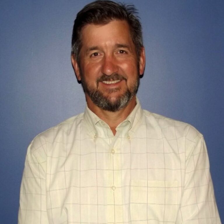 A man with short graying dark hair and a matching beard and mustache smiles against a solid blue background. He wears a pale yellow windowpane-check button-down shirt.