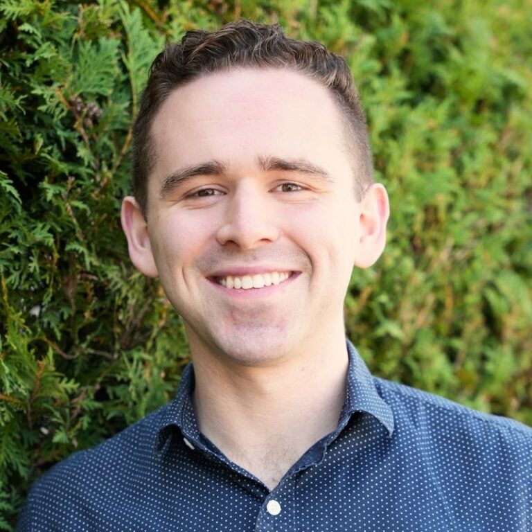 A man with short dark hair styled up and swept back smiles broadly against a backdrop of dense green shrubbery. He wears a navy button-down shirt with a small white polka-dot pattern.