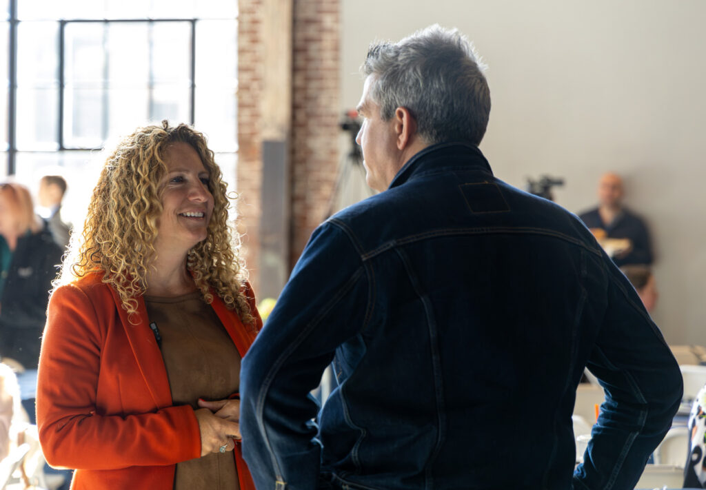 A woman with long curly blonde hair and an orange blazer smiles warmly as she chats with a gray-haired man in a dark denim jacket, who is seen from behind. Soft sunlight streams through a tall window behind her, and a camera on a tripod is visible in the blurred background.