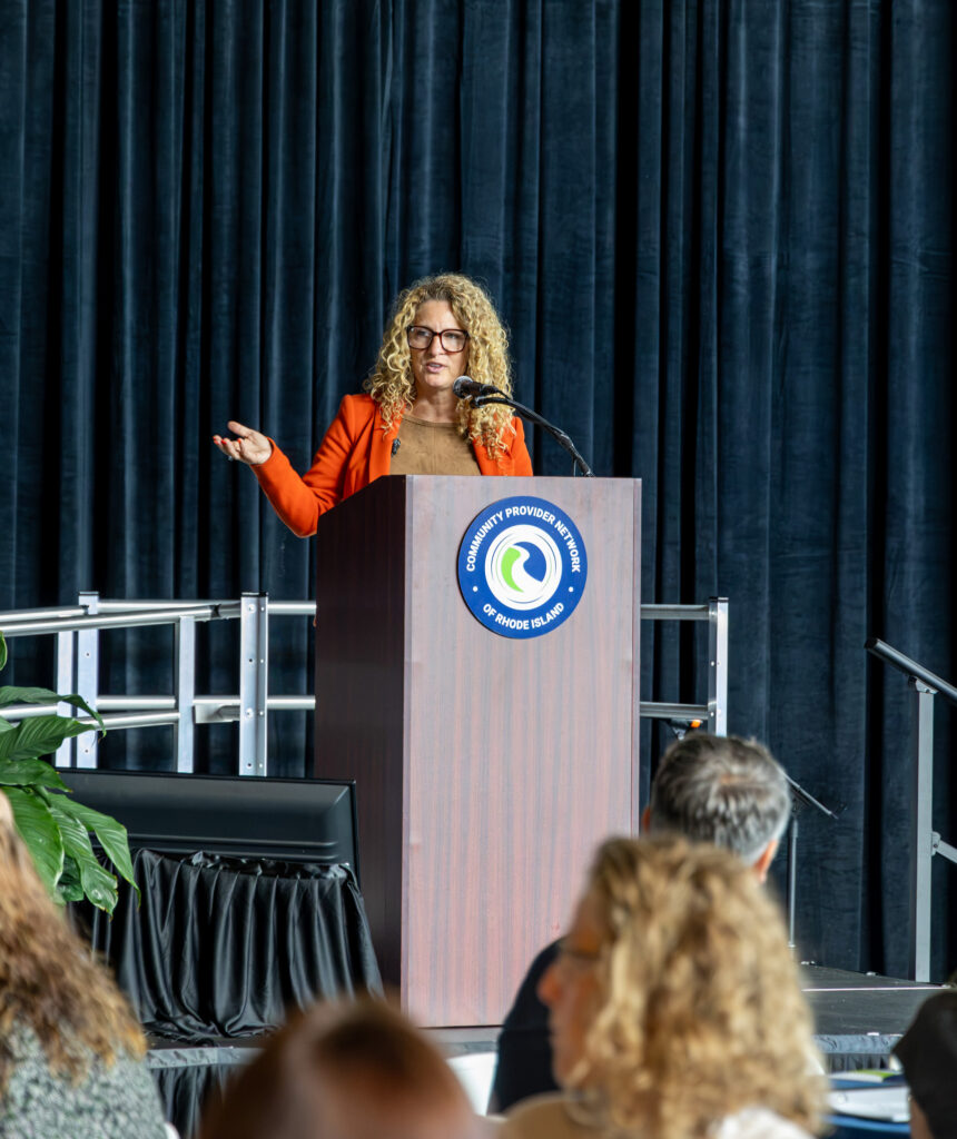 A woman with curly blonde hair, glasses, and an orange blazer speaks at a wooden podium bearing the Community Provider Network of Rhode Island seal, gesturing with one hand. A deep navy velvet curtain hangs behind her, and the backs of audience members' heads are softly blurred in the foreground.