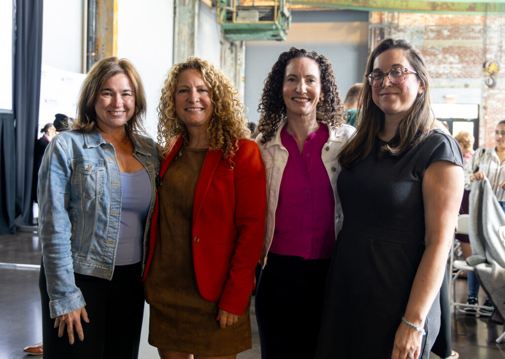 Four smiling women pose shoulder to shoulder inside the brick-walled event hall. From left, they wear a light denim jacket, an orange-red blazer over a brown dress, a white jacket over a magenta blouse, and a dark gray dress with tortoiseshell glasses. Other guests mingle in the background.