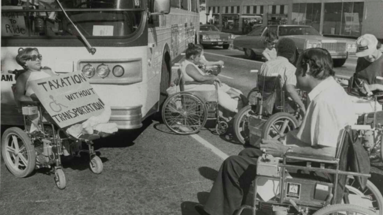 A group of wheelchair users blocks a city bus in the middle of a street during a protest, positioned directly in front of and beside the bus to prevent it from moving. One woman in the foreground holds a large handwritten sign reading Taxation Without Transportation with a wheelchair symbol on it, while several other protesters sit nearby in a line facing the bus and oncoming traffic. Parked and passing cars line the street in the background, with storefronts visible further back.
