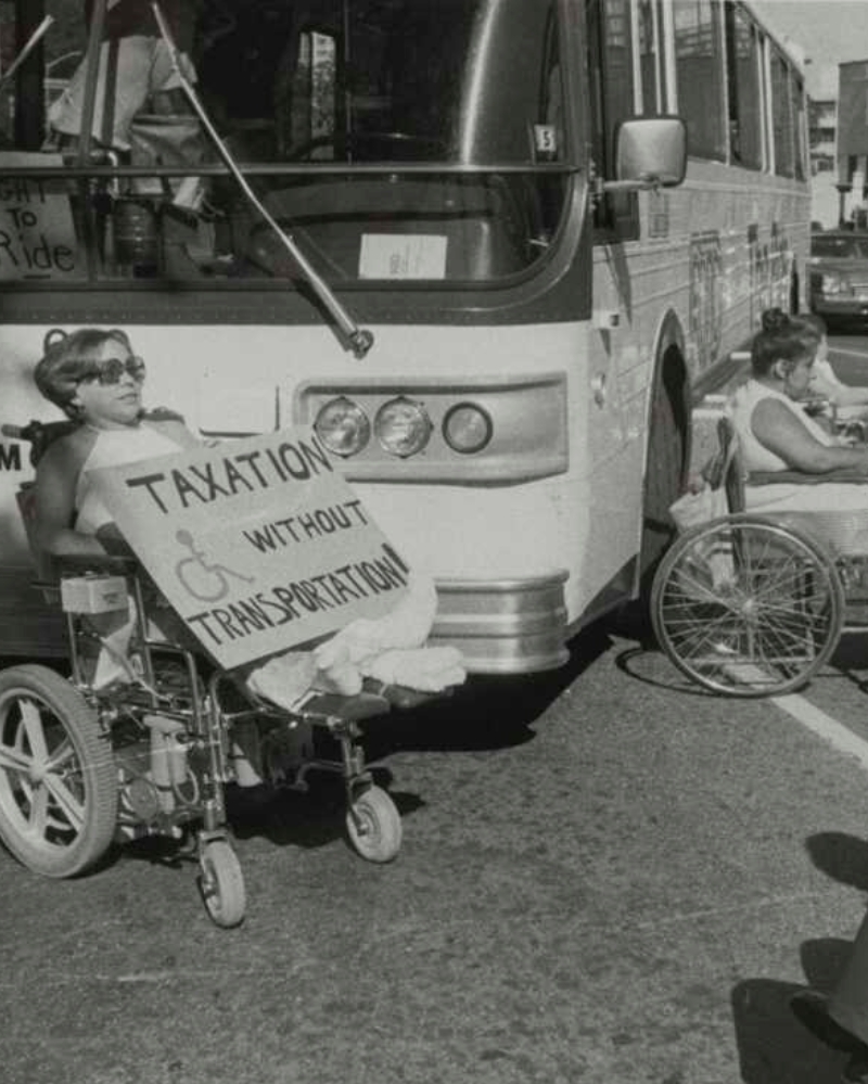 A protester in a wheelchair sits directly in front of the grille of a public transit bus, blocking its path, holding a large handwritten sign reading Taxation Without Transportation with a wheelchair symbol drawn on it. Another wheelchair user is partially visible to the right near the bus door. The black-and-white photo captures a street-level direct-action protest scene.