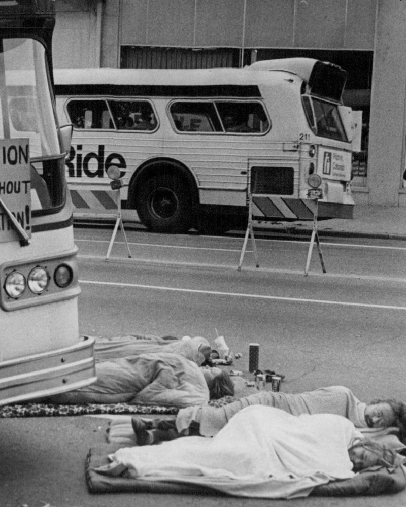 Two protesters lie wrapped in blankets on the pavement directly beneath the front bumper of a transit bus marked Lowry AFB, staging a die-in or lie-in demonstration to block the bus from moving. A handwritten sign reading Taxation Without Transportation is taped to the bus windshield above them. A second bus reading Ride is visible across the street, along with a cooler and gas can sitting on the ground in the foreground.