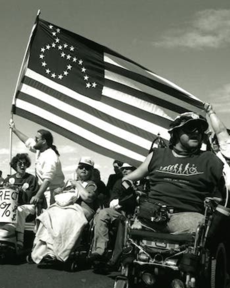 A row of protesters in wheelchairs and scooters move together outdoors, with one man standing and holding up a large flag whose canton has stars arranged in a circular pattern instead of the usual grid. A sign reading Redirect 25% is visible among the group, and participants appear to be chanting or calling out. The wide-open pavement and sky suggest an outdoor rally or march setting.