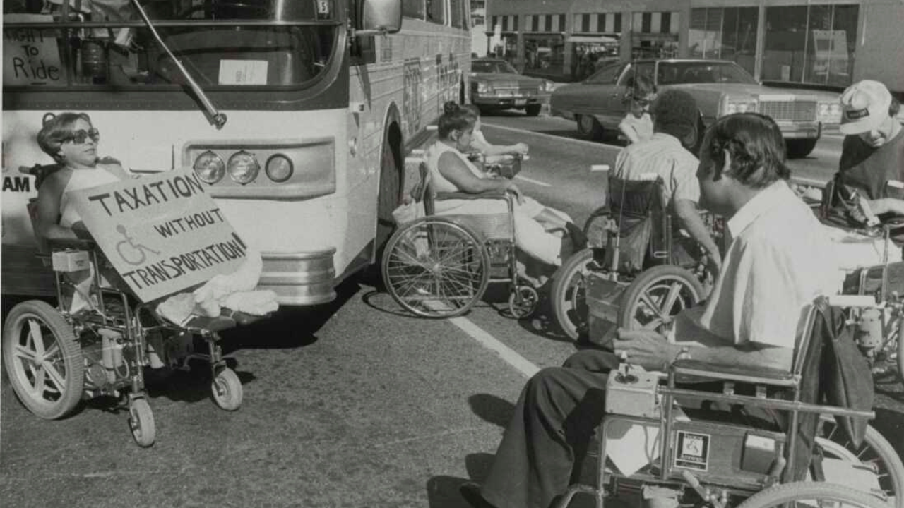 A group of wheelchair users blocks a city bus in the middle of a street during a protest, positioned directly in front of and beside the bus to prevent it from moving. One woman in the foreground holds a large handwritten sign reading Taxation Without Transportation with a wheelchair symbol on it, while several other protesters sit nearby in a line facing the bus and oncoming traffic. Parked and passing cars line the street in the background, with storefronts visible further back.