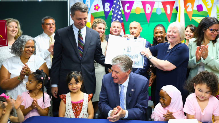 A man in a navy suit sits at a table signing a document while smiling up at a man in a gray suit holding up a clipboard with the signed paperwork. They're surrounded by a large group of cheering, applauding adults and several young children in front of a colorful classroom-themed banner with letters, numbers, and an American flag.