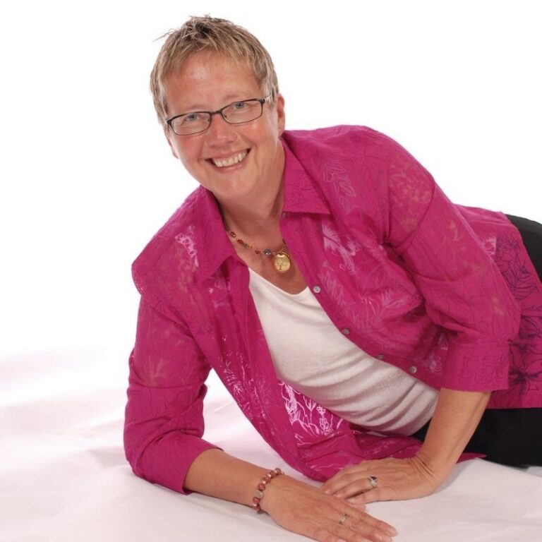 A woman with short blonde hair and rectangular glasses smiles broadly while leaning forward on her forearms and knees against a plain white studio background. She wears a magenta button-down blouse with a subtle floral pattern layered over a white top, along with a beaded bracelet, a pendant necklace, and rings.