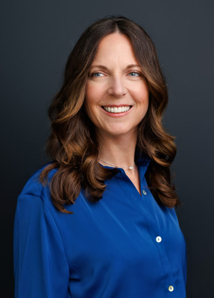 A woman with long wavy brown hair smiles broadly at the camera, wearing a royal blue button-down shirt and a delicate silver necklace, photographed against a dark charcoal studio backdrop.