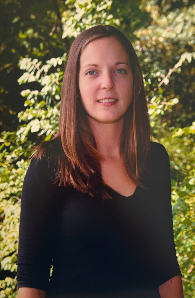 A younger woman with long straight brown hair stands outdoors in front of dense green foliage, wearing a black long-sleeve top and giving a slight, closed smile.