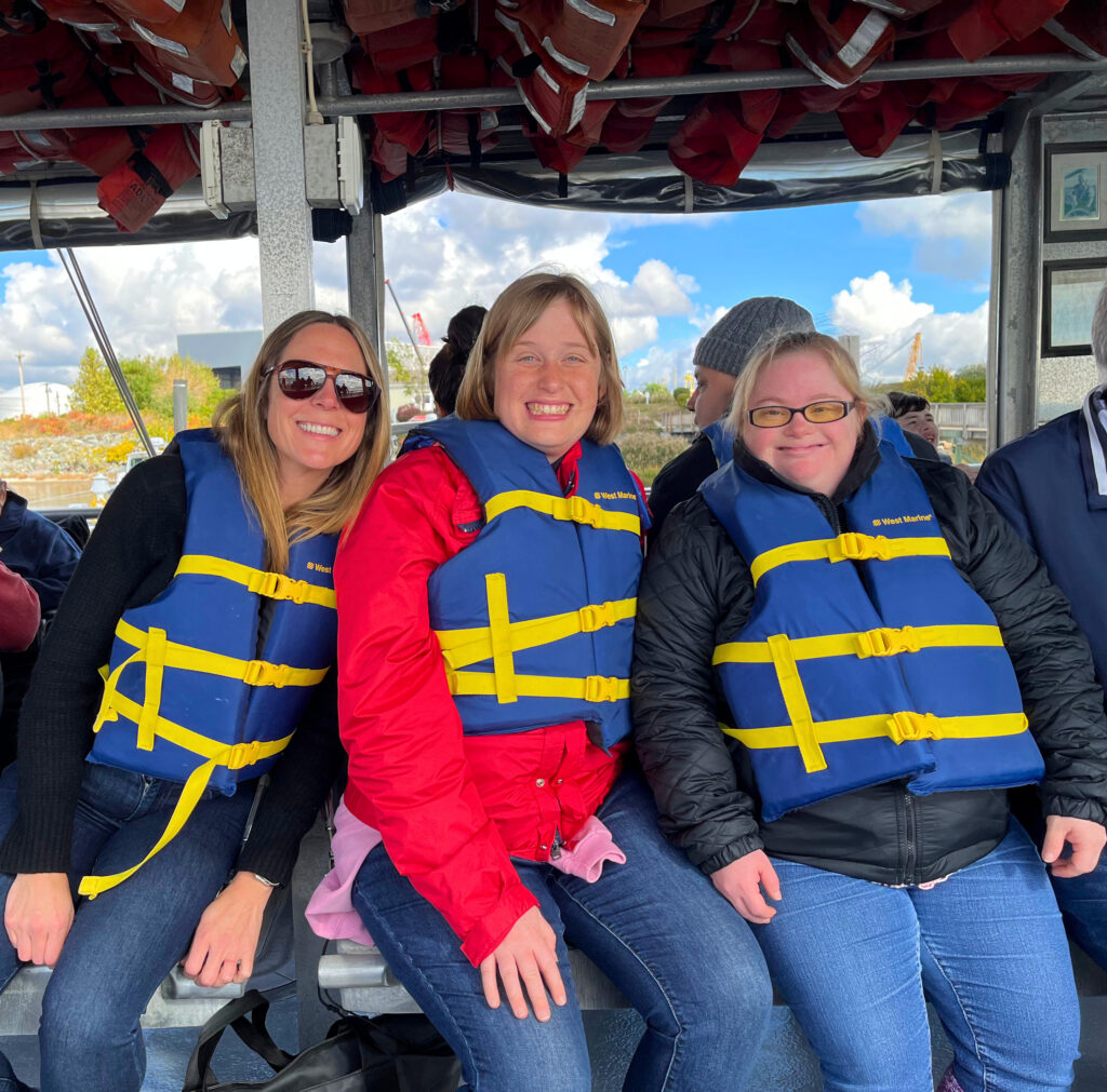 Three women sit together under the canopy of a boat, all wearing blue life jackets with yellow straps over their jackets. The woman on the left wears sunglasses, the woman in the middle wears a red jacket and smiles broadly, and the woman on the right wears glasses with tinted lenses, with a cloudy sky and shoreline visible behind them.