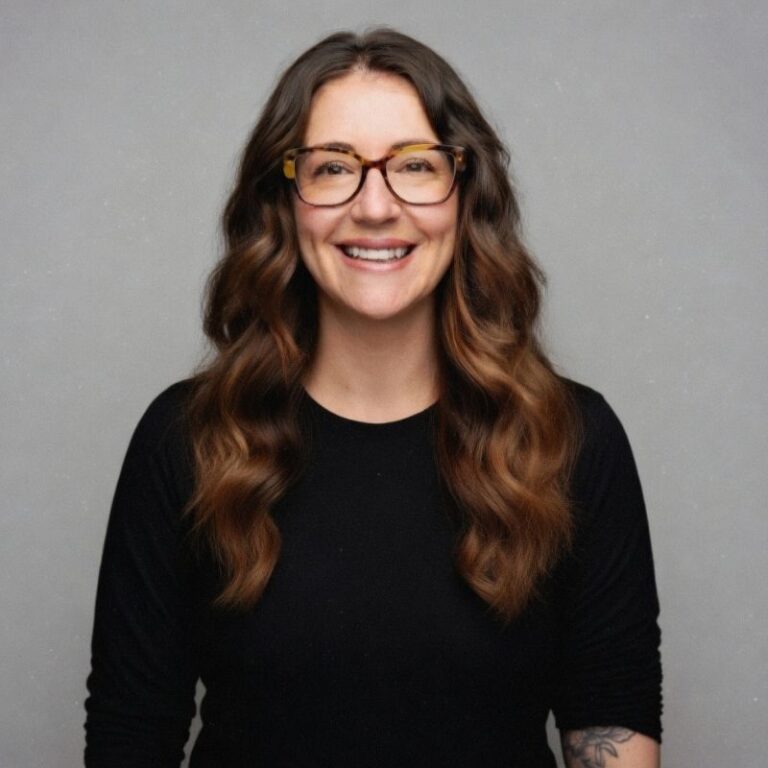 A woman with long wavy auburn hair and tortoiseshell glasses smiles broadly against a plain gray background. She wears a black long-sleeve top, with a visible tattoo on her forearm.