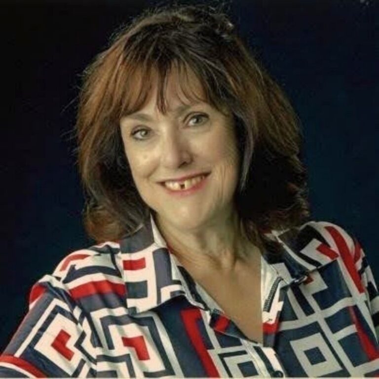 A woman with reddish-brown hair and side-swept bangs smiles against a dark navy background. She wears a bold geometric-print blouse in navy, red, and white with a zip-front collar.