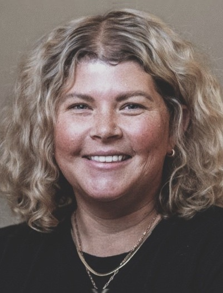 A close-up headshot of a smiling woman facing the camera. She has shoulder-length, naturally curly blonde hair with darker roots and soft waves framing her face. Her hair is parted slightly off center. She has light-colored eyes, full cheeks, and a warm, friendly expression. Her skin has a natural texture with visible freckles across her cheeks and nose. She is wearing small hoop earrings and layered gold necklaces over a black top. The background is a neutral, softly blurred beige tone, keeping the focus on her face. The lighting is even, creating a professional yet approachable portrait style.