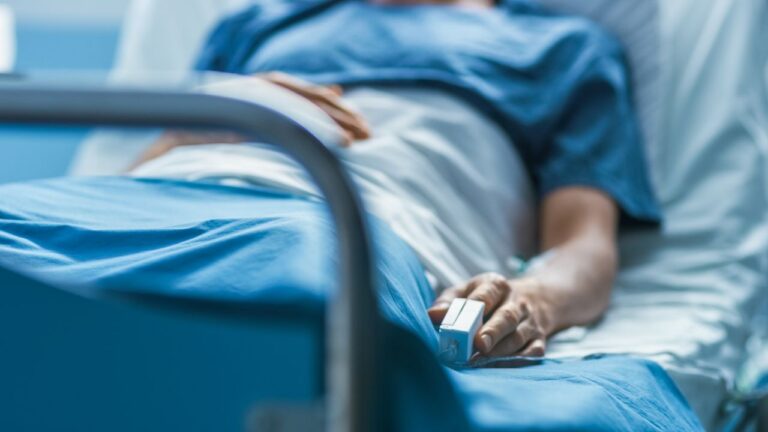 A close-up, softly focused shot of a patient lying in a hospital bed, seen from the side. Their hand rests on the blue blanket with a pulse oximeter clipped to a fingertip, and an IV line is visible near the wrist. The metal bed rail is in sharp focus in the foreground, with the patient and blue-toned hospital linens blurred behind it, giving the image a somber, clinical mood.