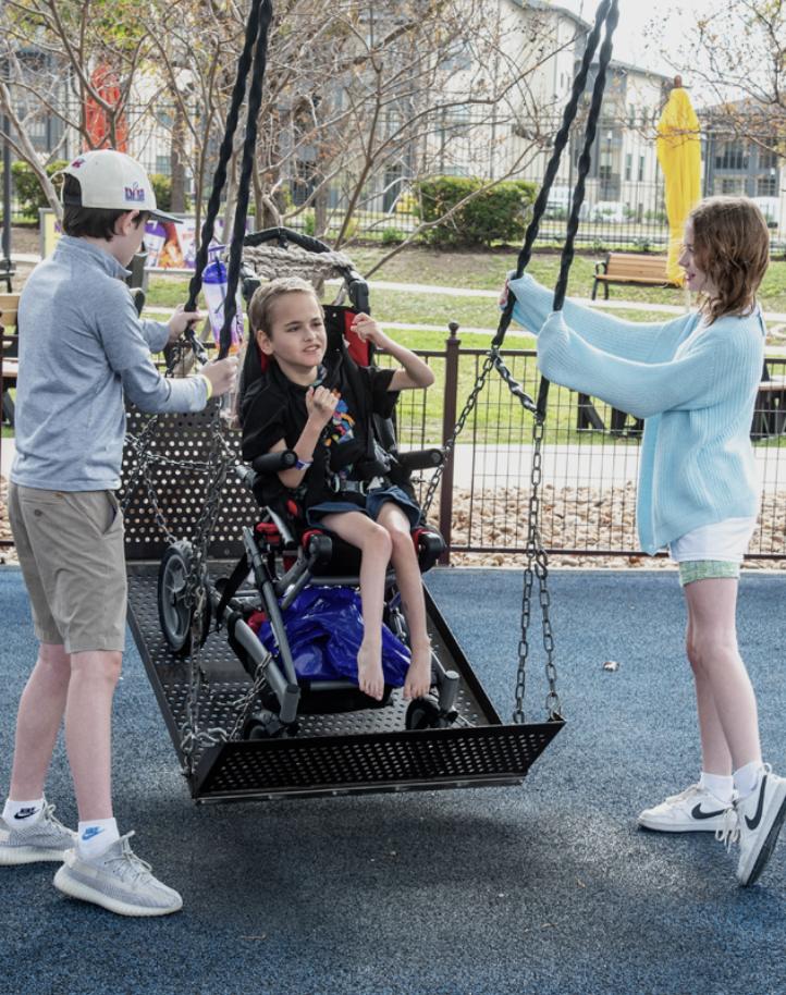 A young child sits secured in a supportive wheelchair that is positioned on an accessible playground swing platform. The swing is a large, black metal platform suspended by four thick chains. The child is barefoot and wearing a black T-shirt and shorts, with a supportive harness visible around their torso. A blue bag is tucked beneath the wheelchair seat. The child’s hands are raised slightly, and their facial expression appears engaged and focused.Two other children stand on either side of the swing, each holding one of the chains. On the left, a child wearing a light gray long-sleeve shirt, khaki shorts, white sneakers, and a light-colored baseball cap faces the swing while steadying it. On the right, another child wearing a light blue oversized sweater, light green shorts, white socks, and white Nike sneakers holds the chain and looks toward the child on the swing.The playground surface is a blue rubberized safety material. In the background, there is a black metal fence, leafless trees, benches, and a light-colored building. A closed yellow patio umbrella stands near the fence, and fallen leaves are scattered on the ground, suggesting a cool season.