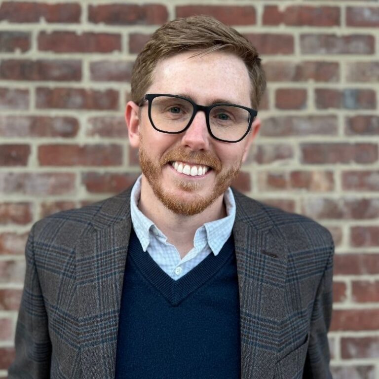 A man with short reddish-brown hair, a matching beard, and dark-framed glasses smiles broadly in front of a red brick wall. He wears a plaid blazer over a navy sweater and light blue collared shirt.