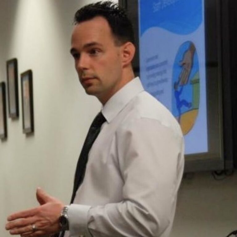 A man with short dark hair stands in profile, speaking and gesturing with one hand while wearing a white button-down shirt and dark tie. Behind him, a framed illustration is projected on a screen, and a few small picture frames hang on a white wall to the left.