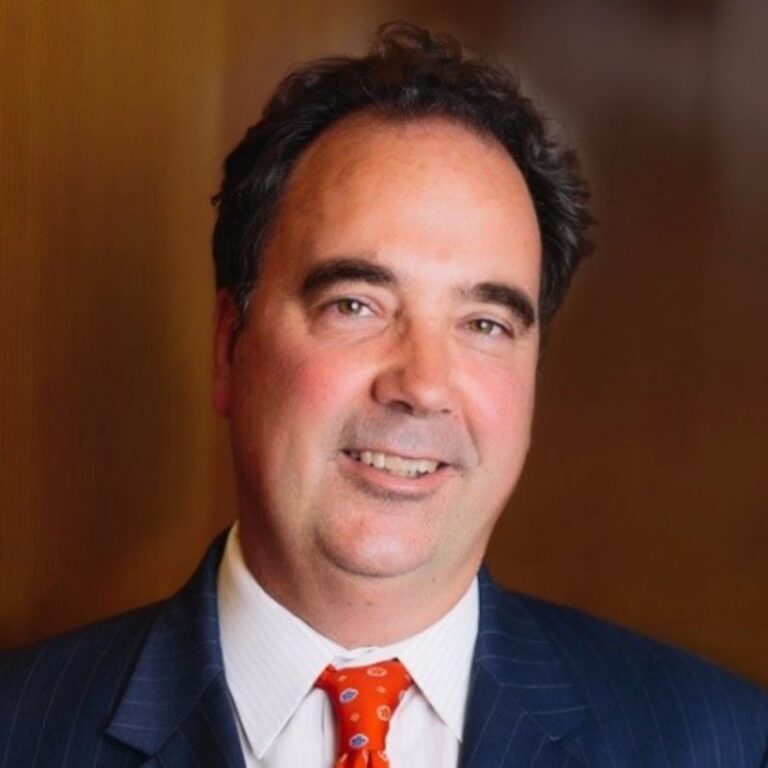 A man with dark hair on the sides smiles warmly against a warm brown wood-paneled background. He wears a navy pinstriped suit with a white shirt and an orange patterned tie.