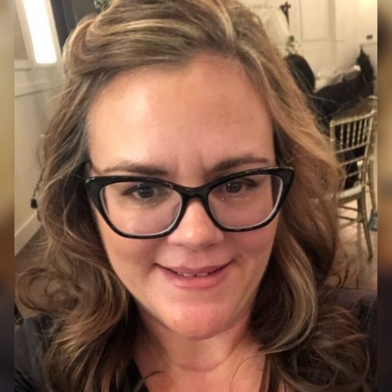 A woman with wavy light brown hair and dark cat-eye glasses smiles gently in a close-up selfie taken indoors, with wooden folding chairs and a wall cross visible in the softly blurred background. She wears a dark top.