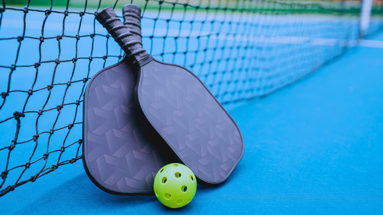 Two dark pickleball paddles with textured, honeycomb-patterned faces and black perforated grips lean crossed against a black net, with a bright green perforated pickleball resting at their base. The scene sits on a blue outdoor court, with the net's white top band and a court post visible blurred in the background.