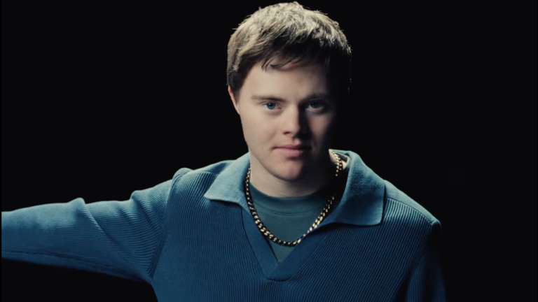 A young person with short brown hair looks directly at the camera against a dark background. They are wearing a blue textured sweater over a matching shirt and a gold chain necklace. One arm is extended out to the side, partially out of frame. The lighting is dramatic, highlighting their face while the background remains black.