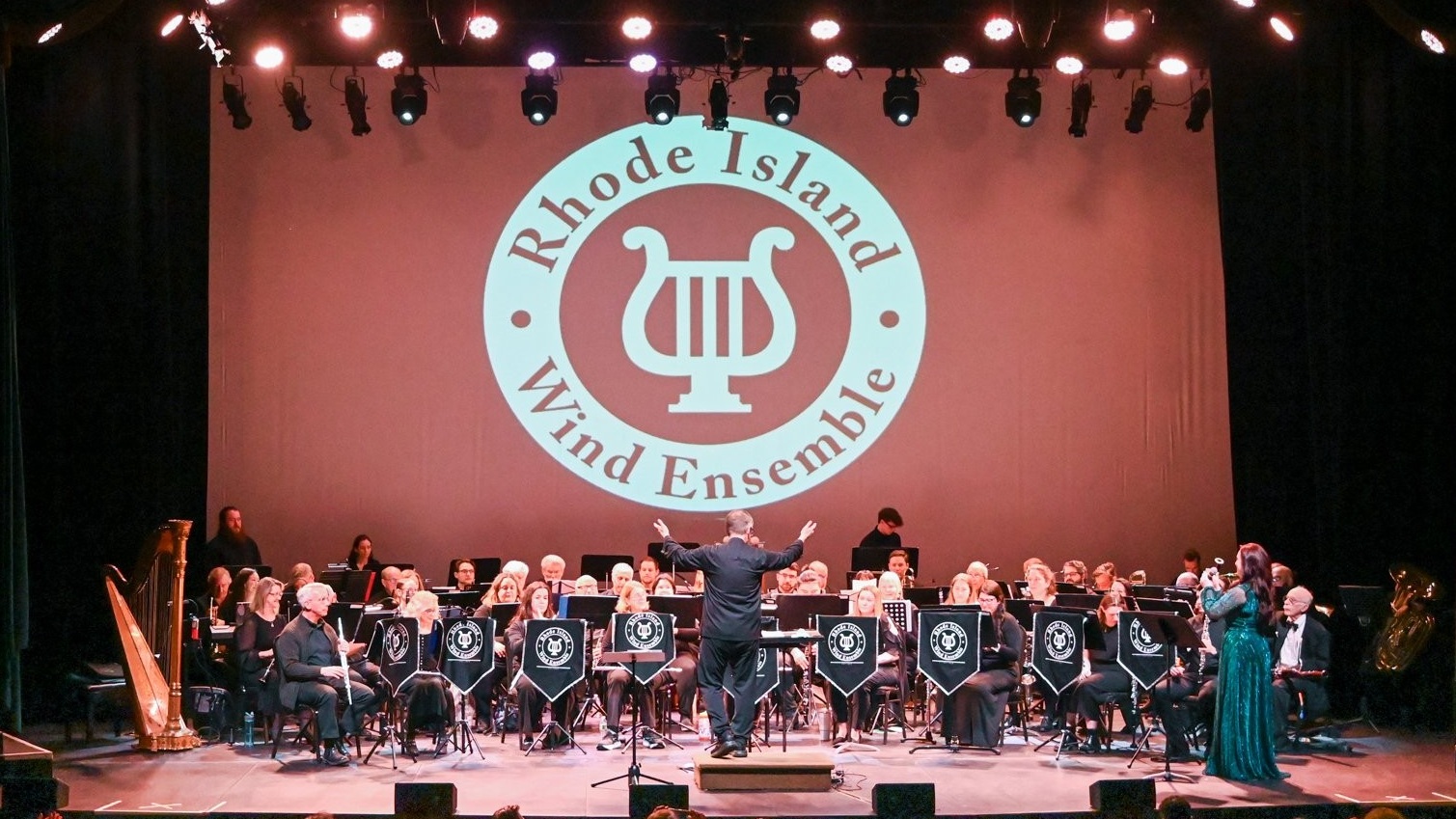 Rhode Island Wind Ensemble performing onstage with musicians seated beneath the ensemble’s logo.
